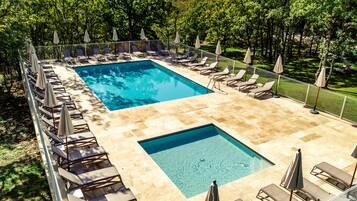 A rectangular pool with two smaller sections, surrounded by lounge chairs and umbrellas, set against a backdrop of trees and a hilly landscape.