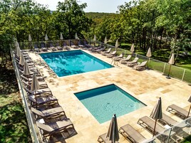 Una piscina rectangular con dos secciones más pequeñas, rodeada de sillas de descanso y sombrillas, con árboles y un paisaje montañoso de fondo.