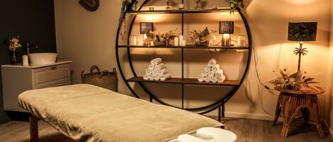 Un cuarto de masajes con un banco de madera, una toalla blanca y un estante con velas y plantas.