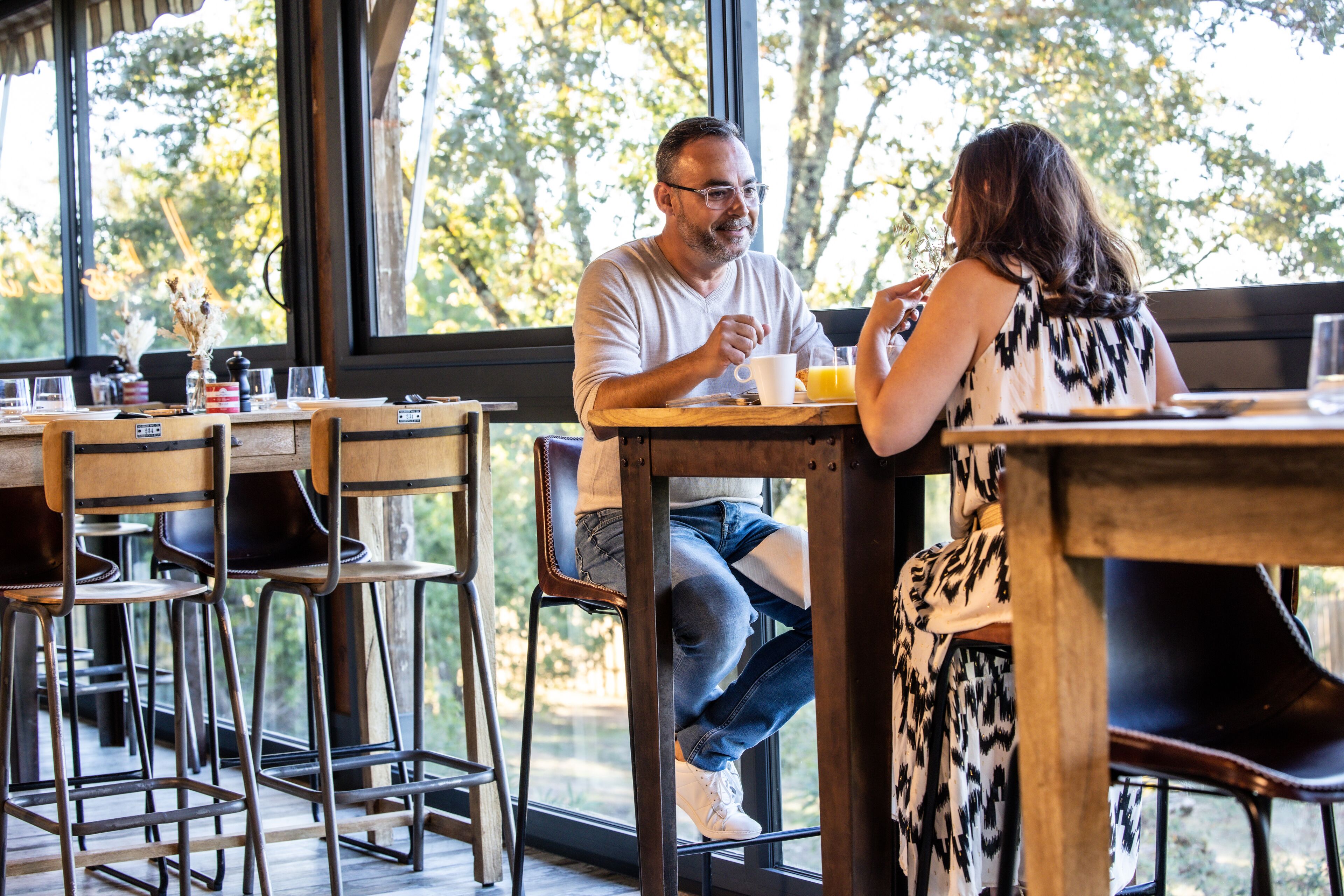 A dining area with wooden tables and chairs, large windows with a view of trees, and a casual atmosphere.