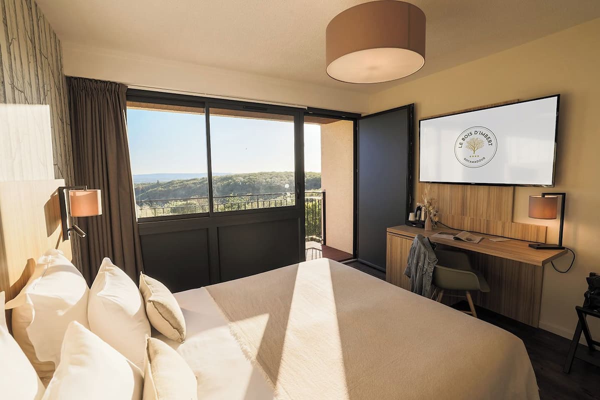 A hotel room with a large bed, a desk, a chair, a TV, and a view of the outdoors.
