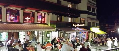 A bustling outdoor dining area with multiple tables and chairs, surrounded by buildings with illuminated signs.