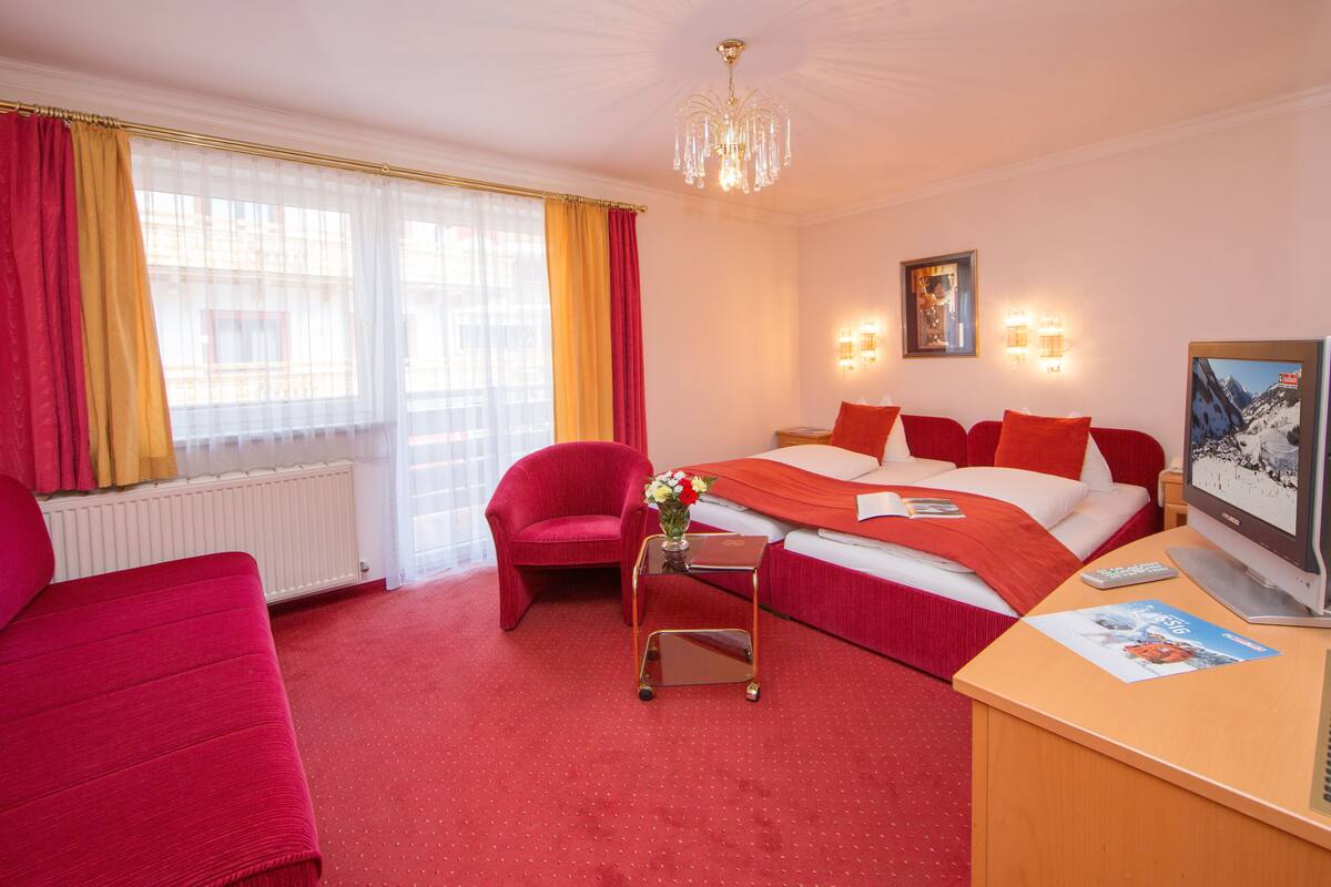 Ein Hotelzimmer mit einem Bett, einem roten Sessel, einem kleinen Tisch mit Blumen in einer Vase, einem Fernseher und einem Fenster mit Vorhängen.