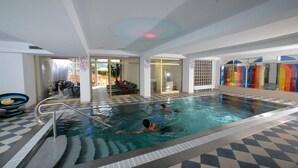 Una piscina interior con dos personas nadando, baldosas de suelo a cuadros y una pared de cristal con vidrieras de colores.