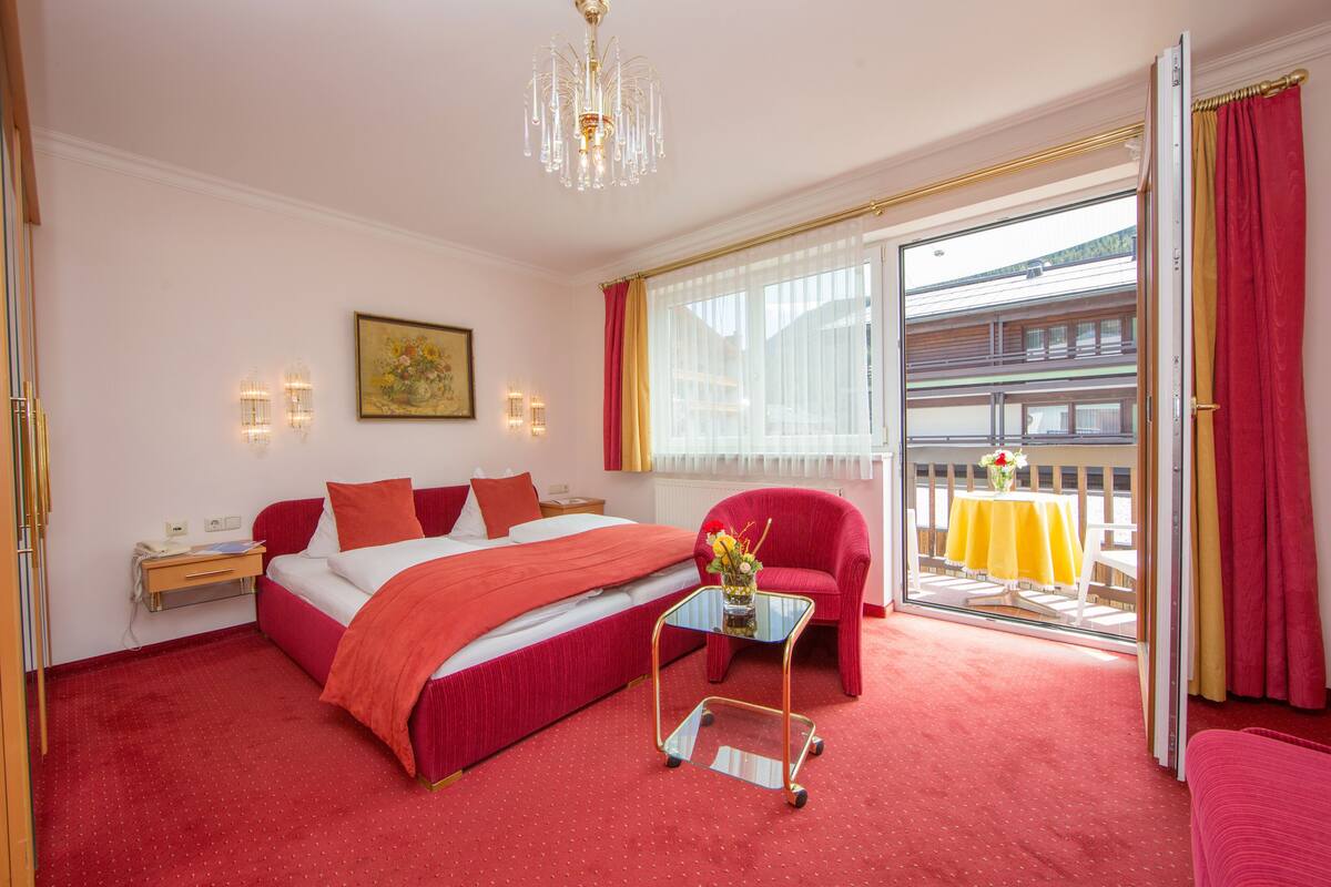 Ein Hotelzimmer mit einem großen Bett, einem roten Sessel, einem kleinen Tisch mit einer Vase und einem Balkon mit einem gelben Tisch.