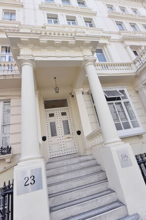 Un edificio con facciata neoclassica, una scalinata che conduce a una porta con decorazioni, e un cartello che indica il numero civico 23.