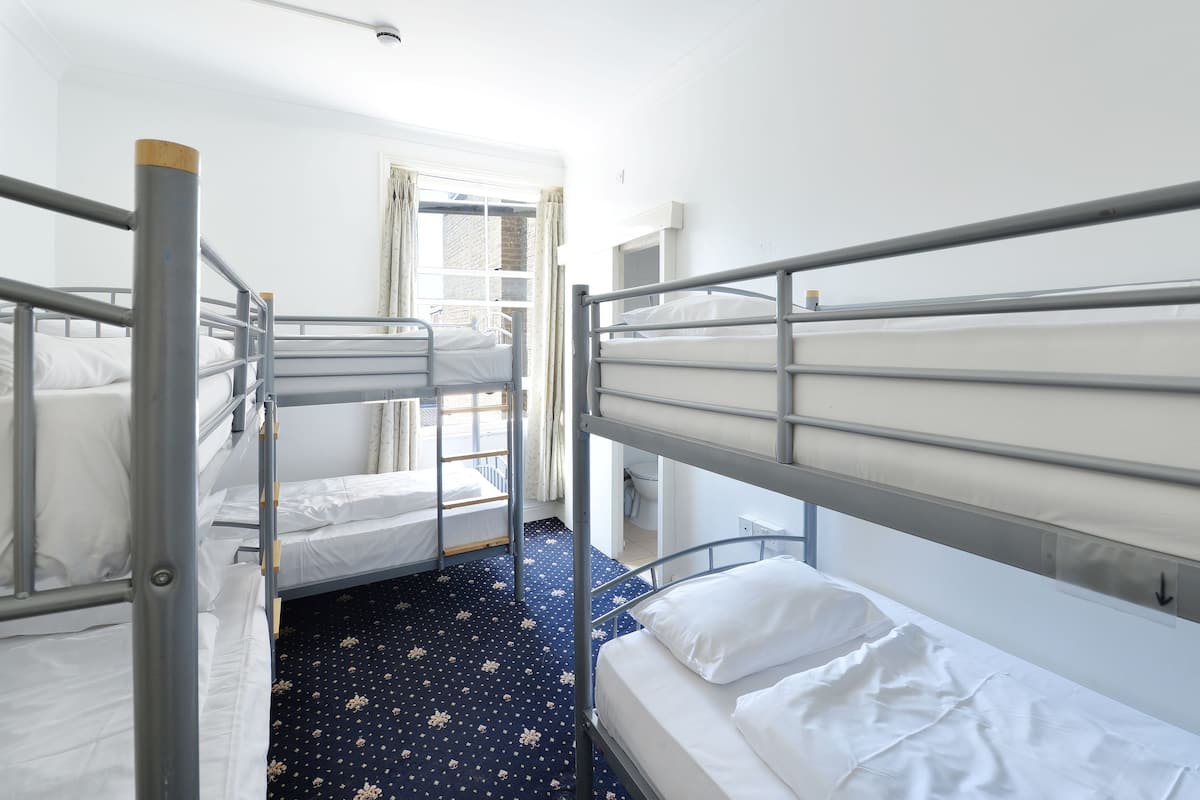 A dormitory room with bunk beds, a window with curtains, and a patterned carpet.