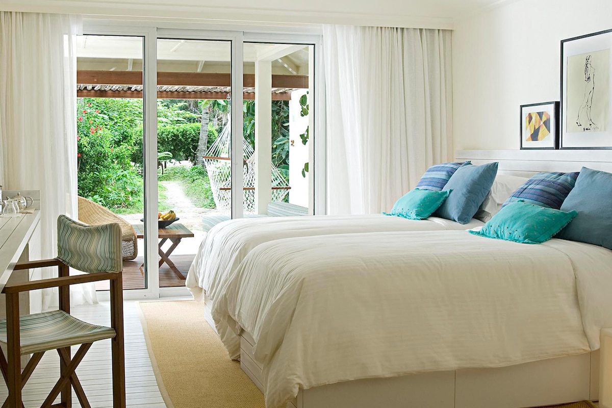 A bedroom with a bed, a desk, a chair, and a television. There is a sliding glass door leading to an outdoor area with greenery.