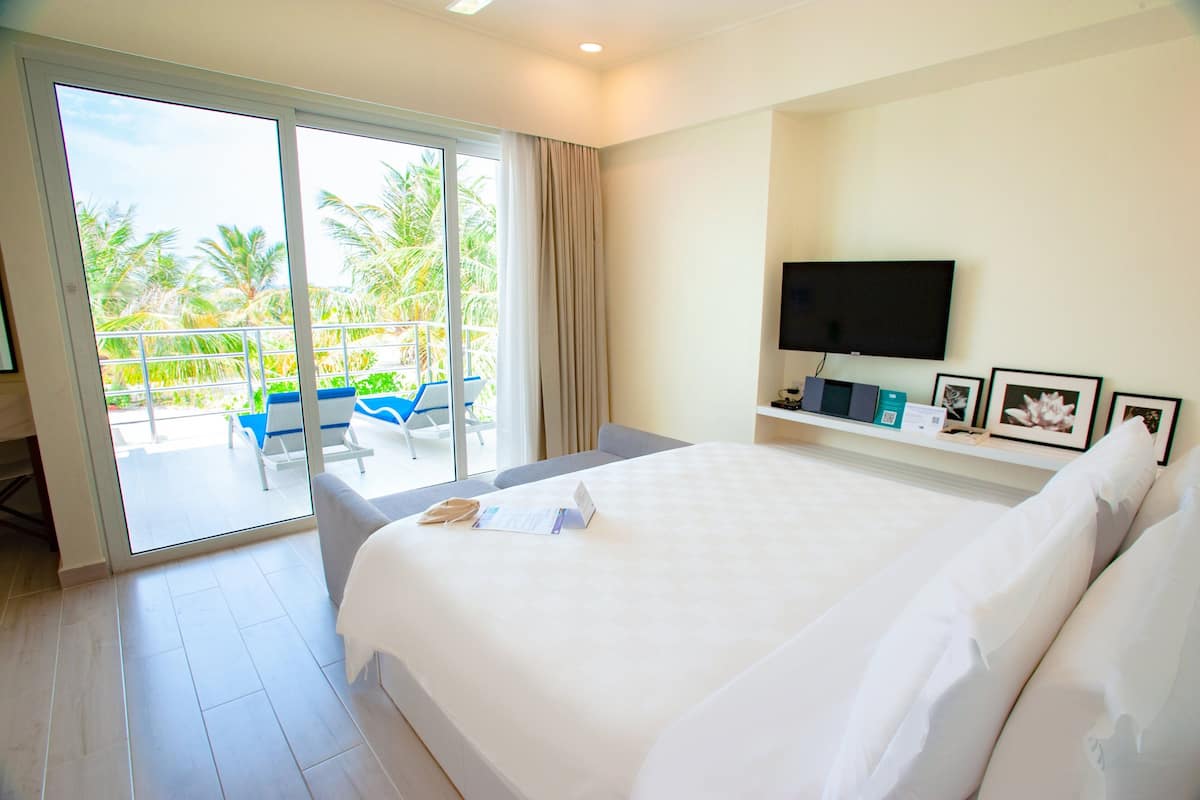 A hotel room with a large bed, a TV, a balcony with a view of palm trees, and a desk with a chair.