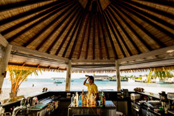 A beachside bar with a thatched roof, a bartender, and a view of the beach and ocean. - Holiday Inn Resort Kandooma Maldives by IHG (Kandooma)