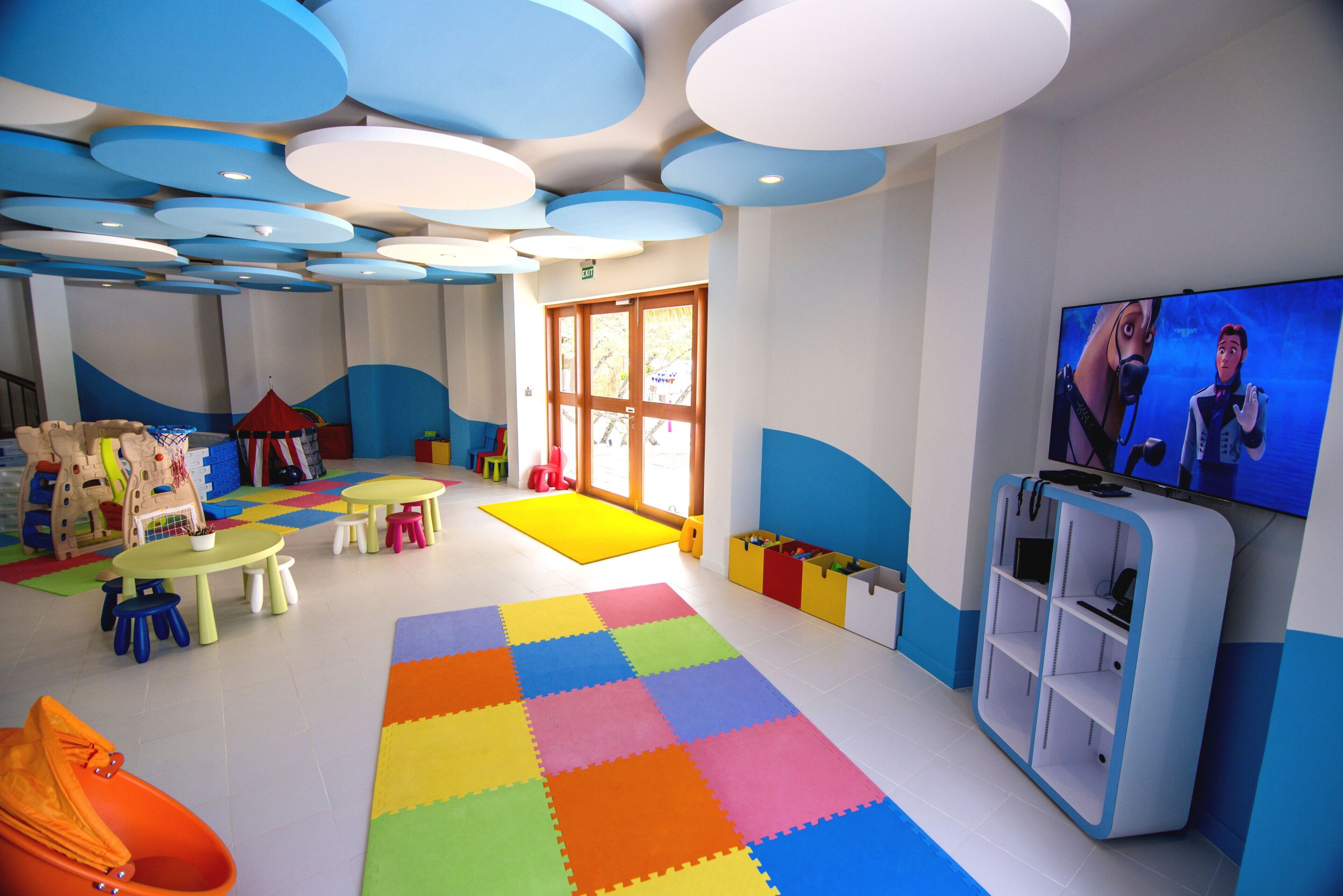 Ein Kinderspielbereich mit farbenfrohem Boden, Tischen, Stühlen und einem Fernseher.