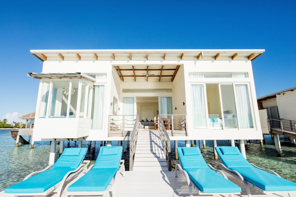 Un bungalow blanc sur pilotis, doté d’un escalier central menant à un espace de vie, entouré d’une eau d’un bleu cristallin et agrémenté de chaises longues bleues.