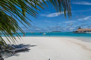 A beach with white sand, clear turquoise water, and boats. There are lounge chairs and a building with a thatched roof.