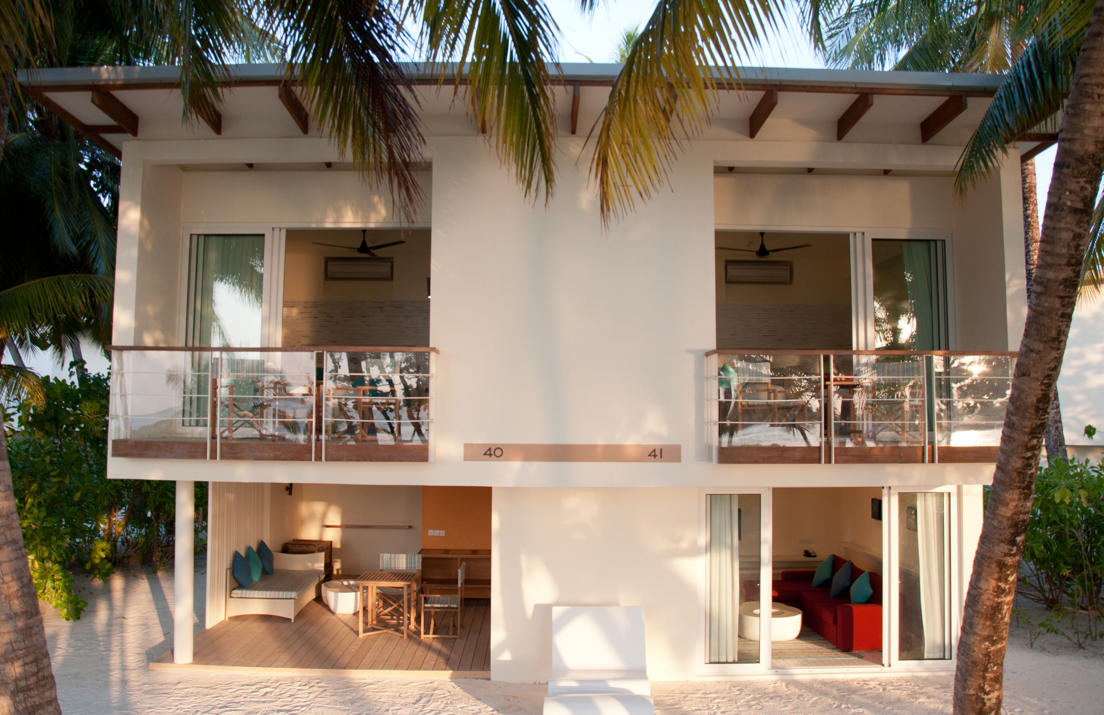 A modern two-story house with a balcony, a patio with outdoor furniture, and a view of the beach.