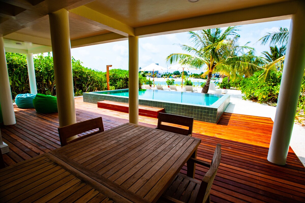 Une terrasse en bois avec une piscine et des chaises longues, entourée de palmiers et de verdure.