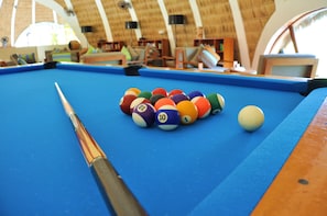 A pool table with a variety of numbered balls and a cue stick, set in a room with wooden furniture and a thatched roof. - Holiday Inn Resort Kandooma Maldives by IHG (Kandooma)