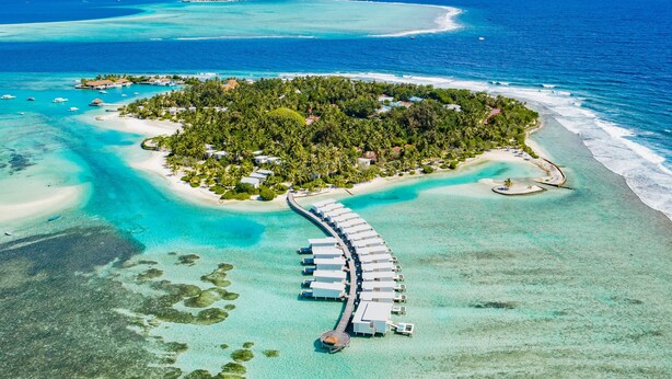 An aerial view of a tropical island resort with overwater bungalows connected by a long pier, surrounded by clear turquoise waters and lush greenery. - Holiday Inn Resort Kandooma Maldives by IHG (Kandooma)
