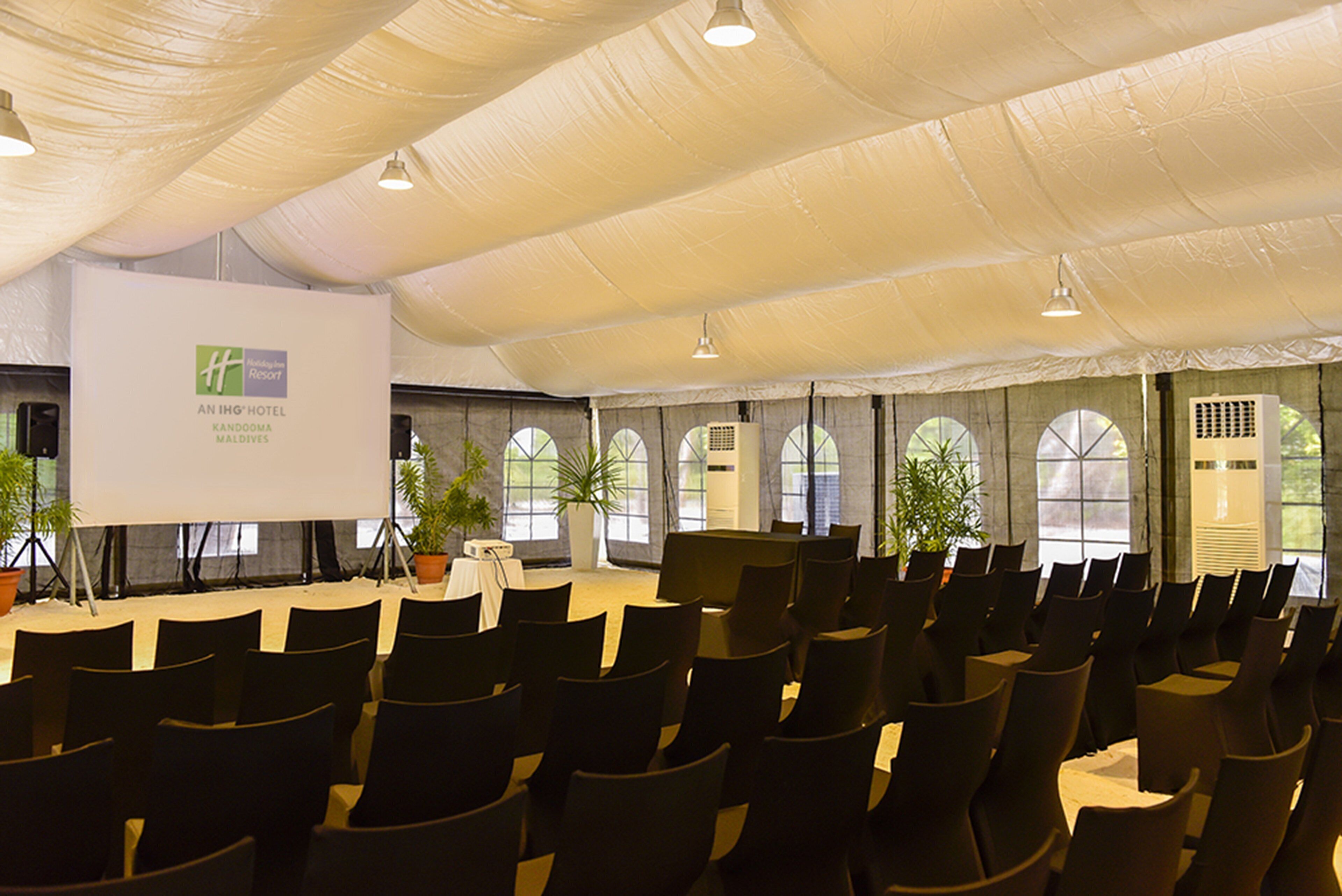 Un salón de conferencias con filas de sillas, una pantalla de proyección y plantas en macetas.