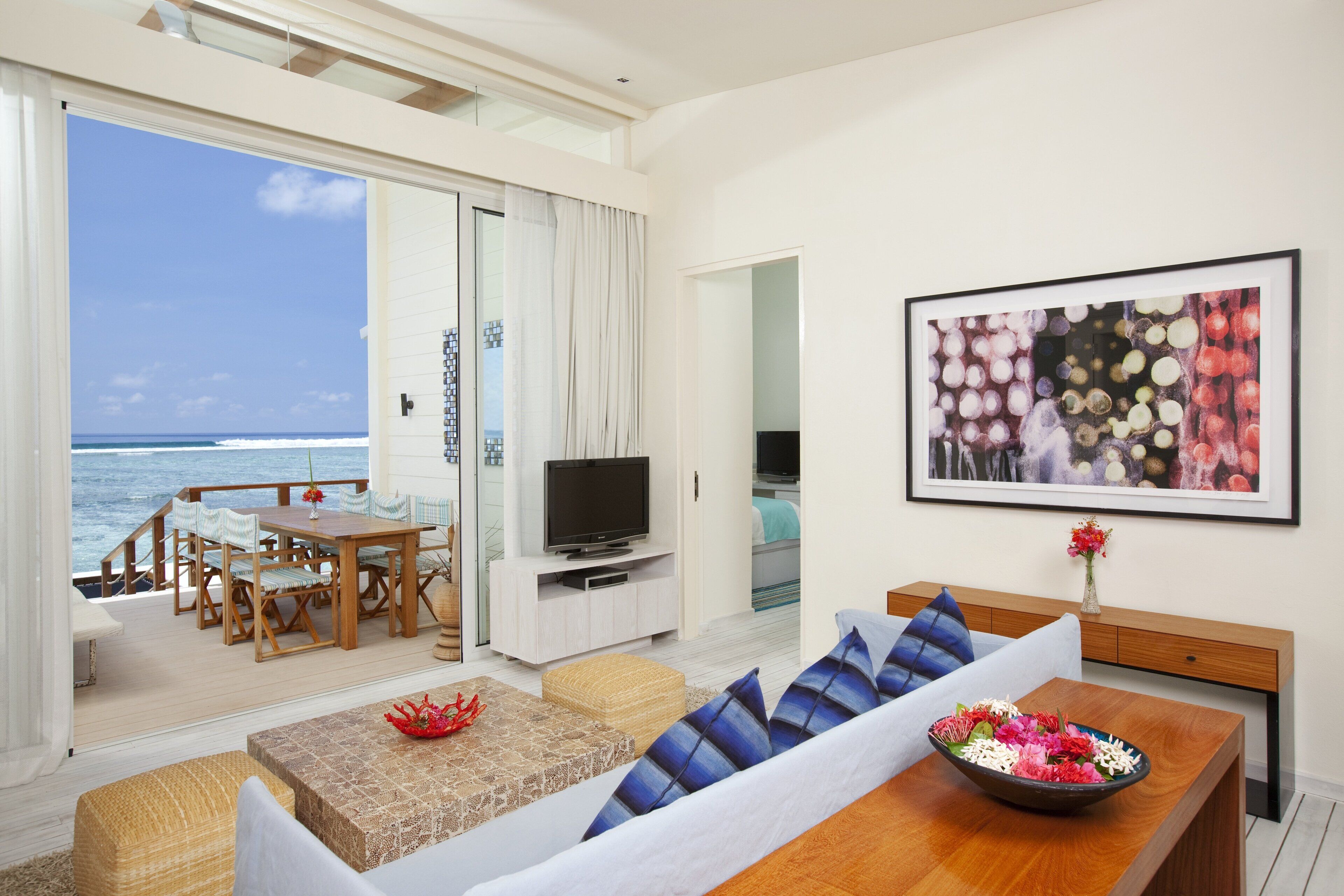 A living room with a TV, a sofa, a dining table, and a view of the ocean.
