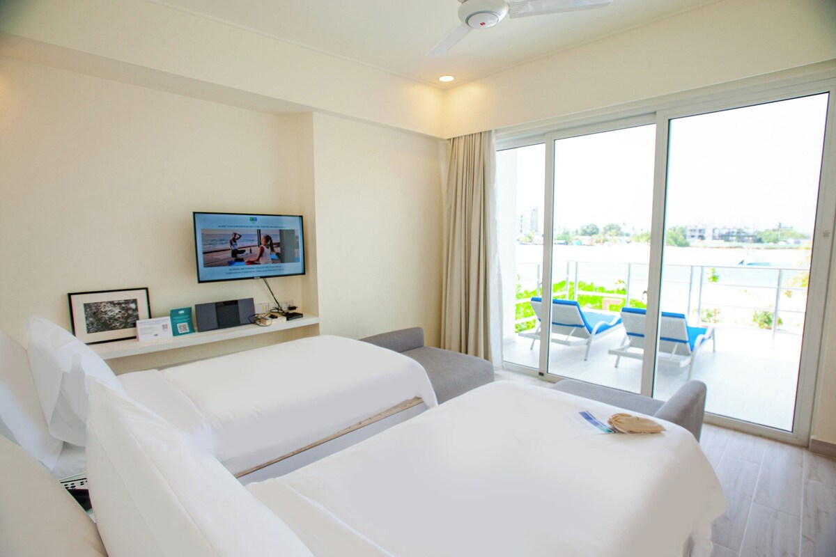 A hotel room with two beds, a TV on the wall, and a balcony with outdoor seating.