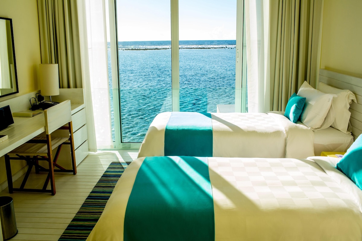 A hotel room with a large window offering a view of the sea, two beds with matching bedding, a desk with a lamp, and a trash bin.