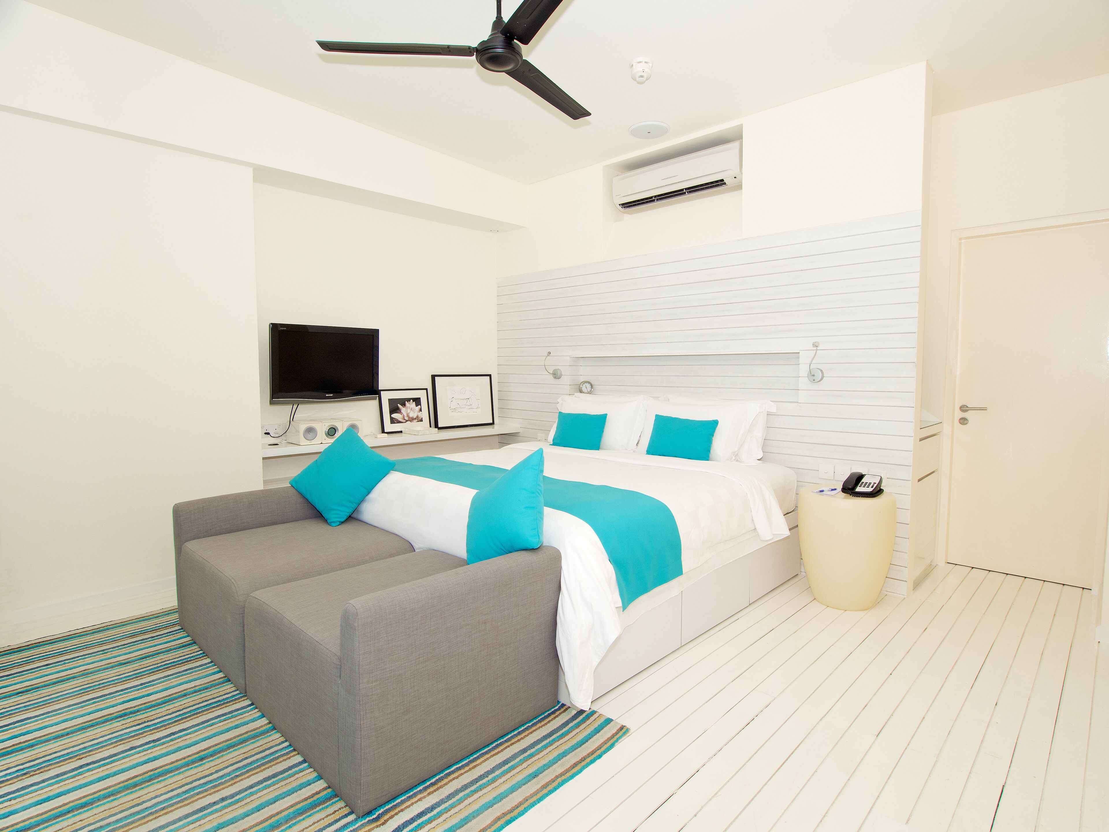 A modern bedroom with a bed, a grey sofa, a television, and a ceiling fan.