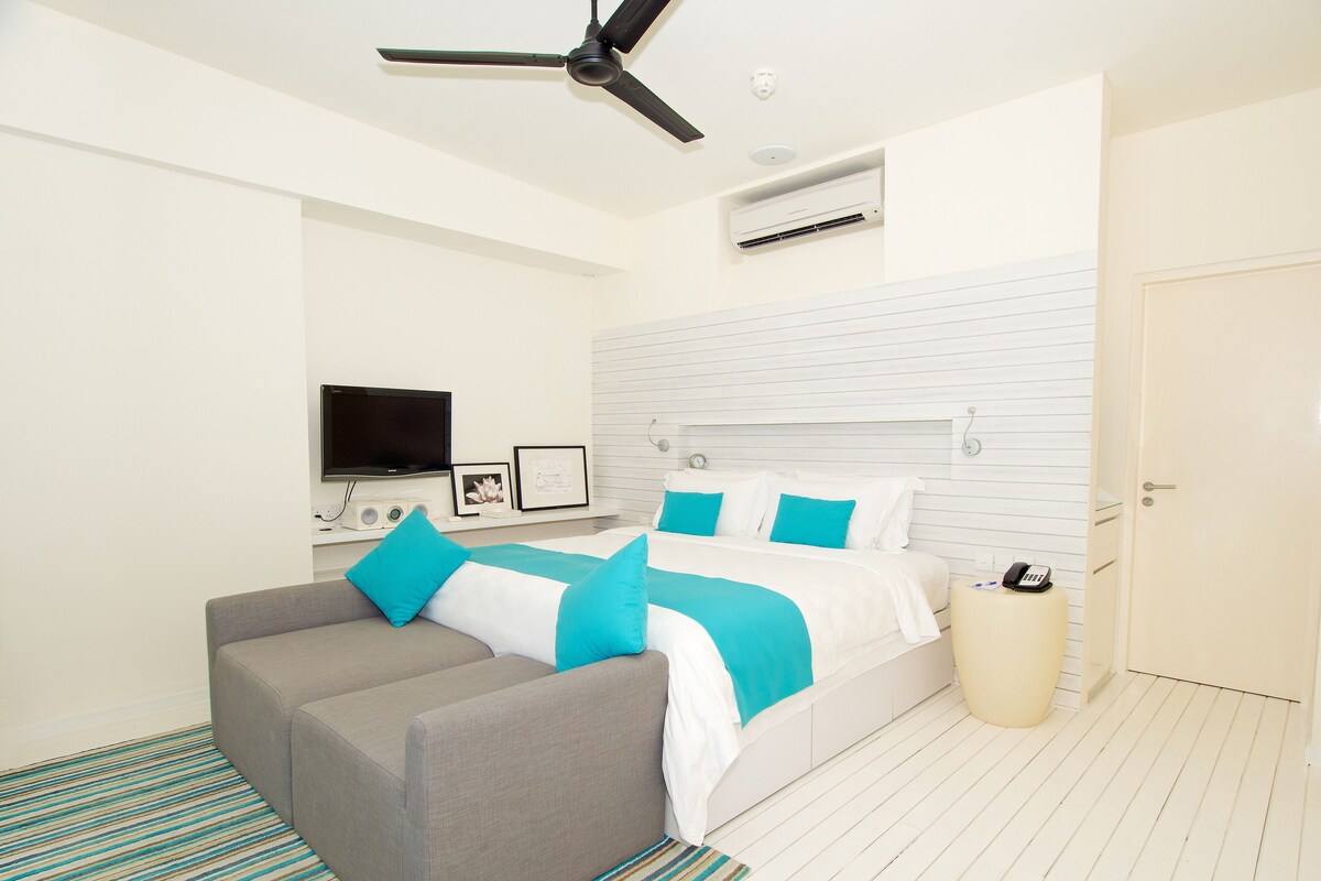 Une chambre à coucher moderne avec un lit, un canapé gris, une télévision et un ventilateur de plafond.