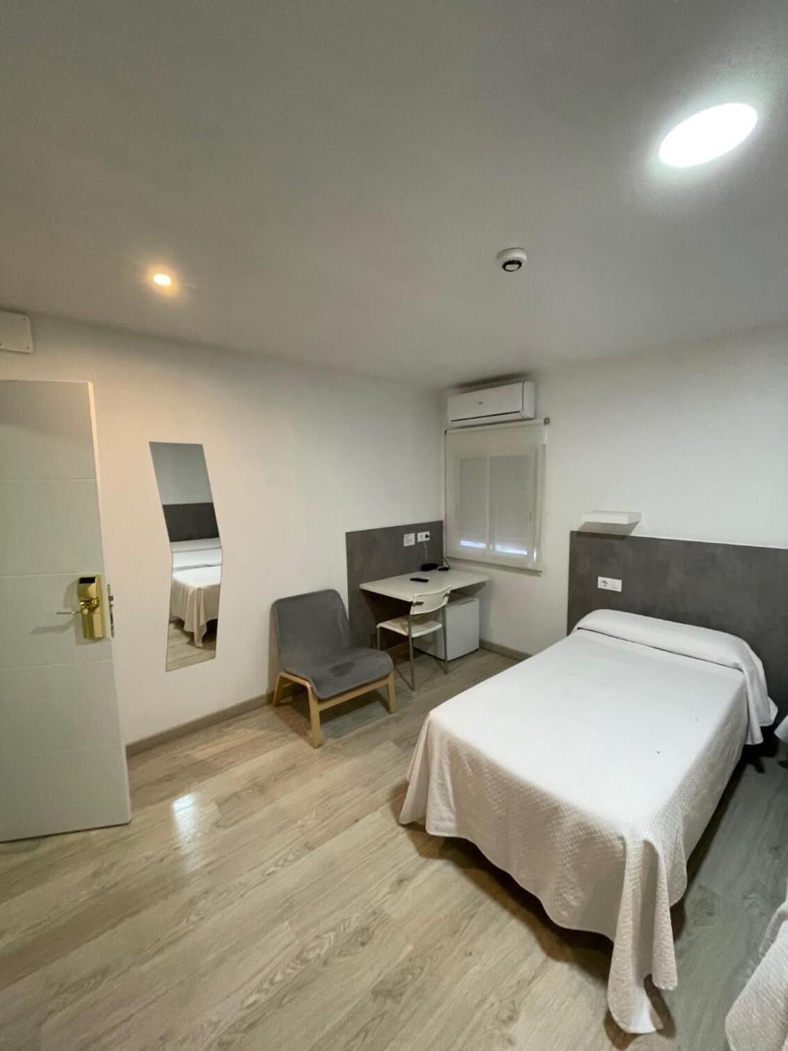 A hotel room with a bed, a desk, a chair, a mirror, and an air conditioning unit.