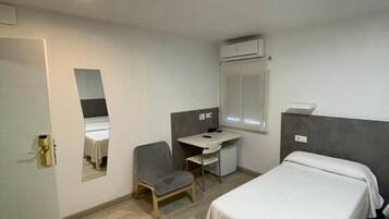 A hotel room with a bed, a desk, a chair, a mirror, and an air conditioning unit.