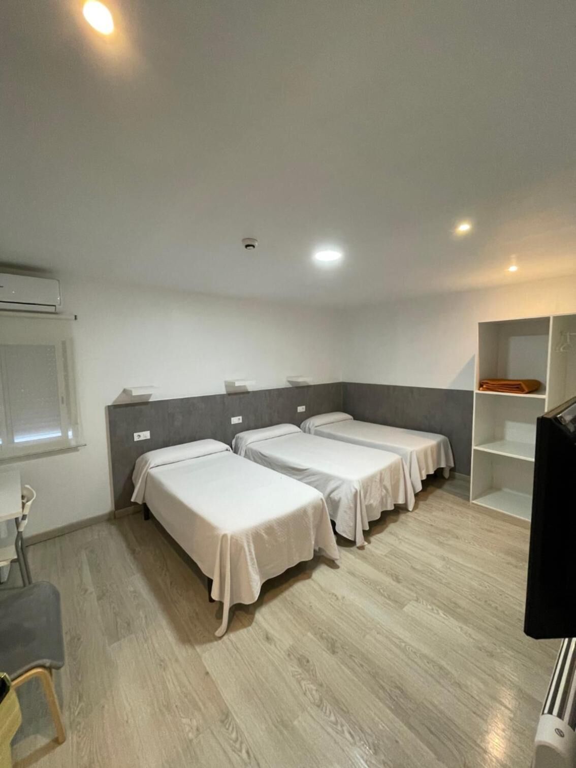 Habitación de hotel con tres camas individuales, un escritorio, una silla, un televisor y una estantería con objetos doblados.