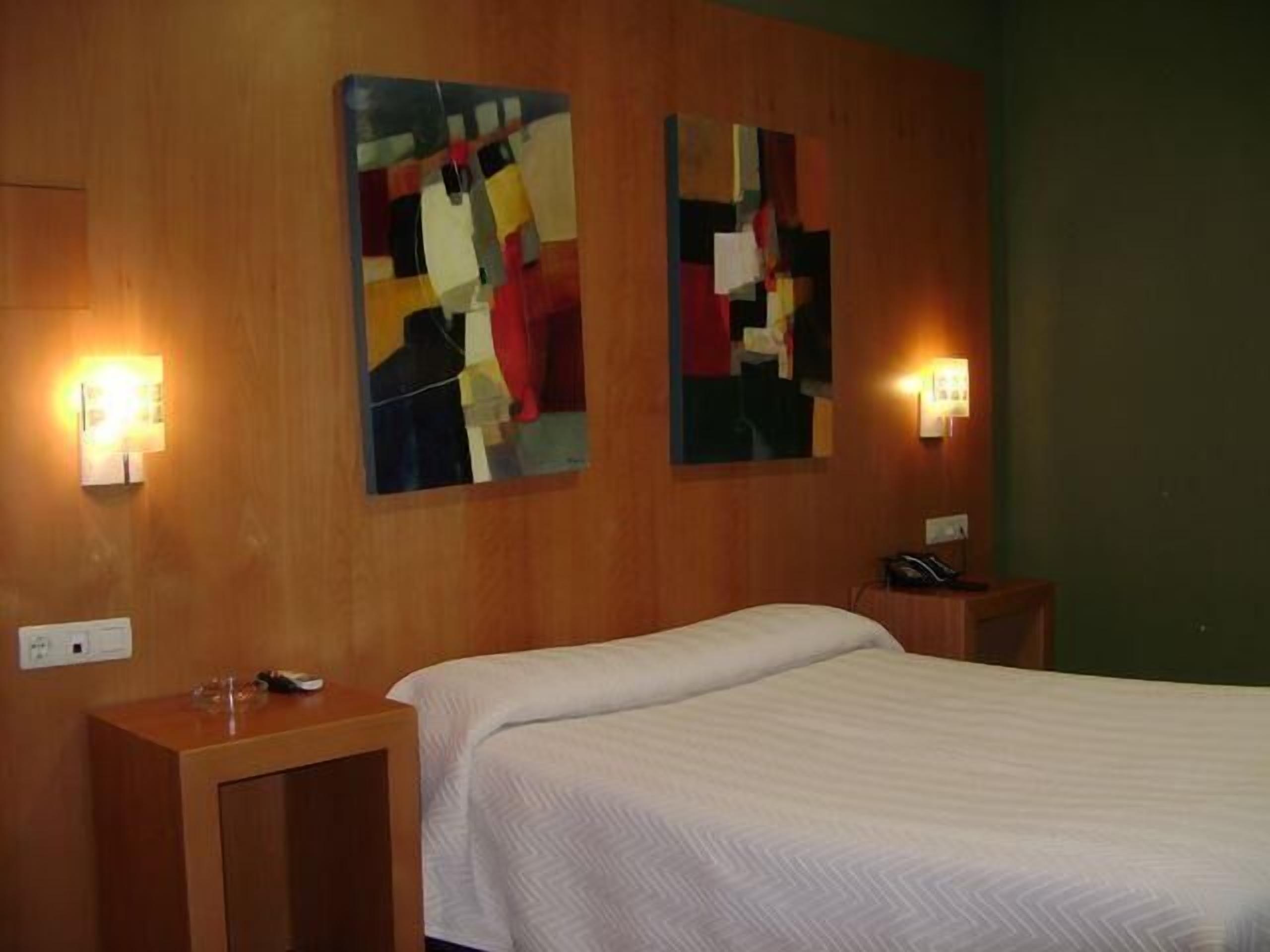 Una habitación de hotel con una cama, dos cuadros colgados en la pared, mesitas de noche y apliques.