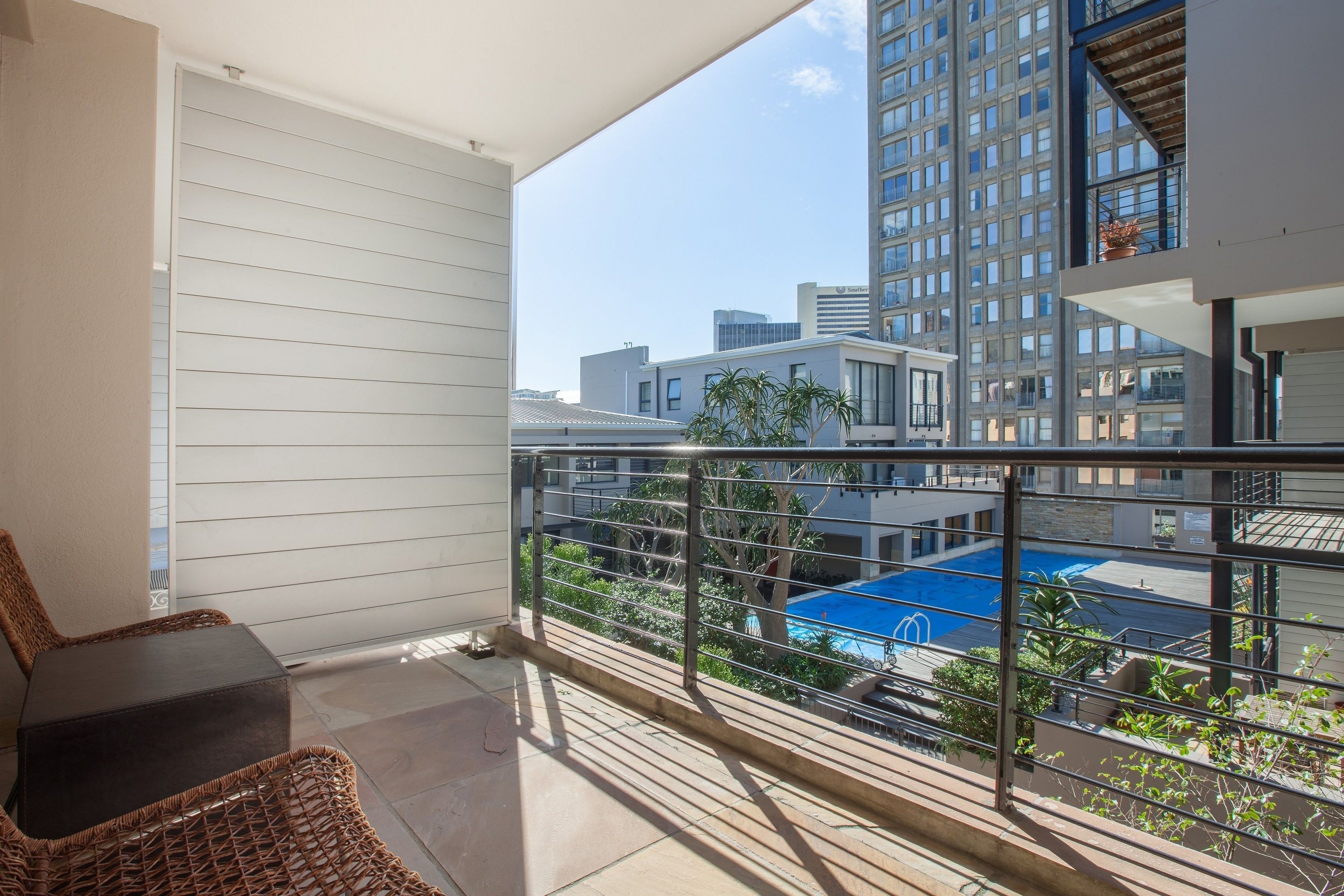 Un balcon avec un fauteuil en osier et une petite table, donnant sur une piscine bleue et les bâtiments environnants.