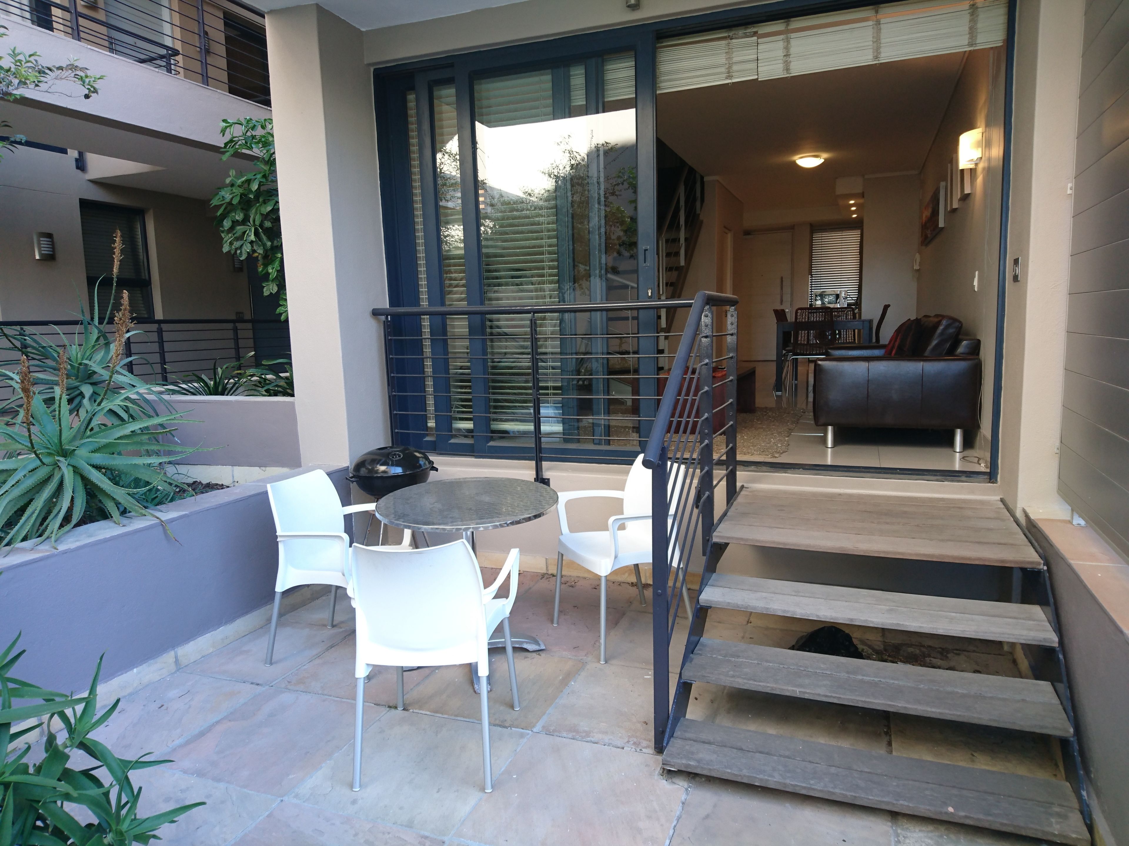 Une terrasse avec une table ronde et deux chaises blanches, un barbecue noir et un escalier menant à un espace intérieur comprenant un canapé et un coin repas.