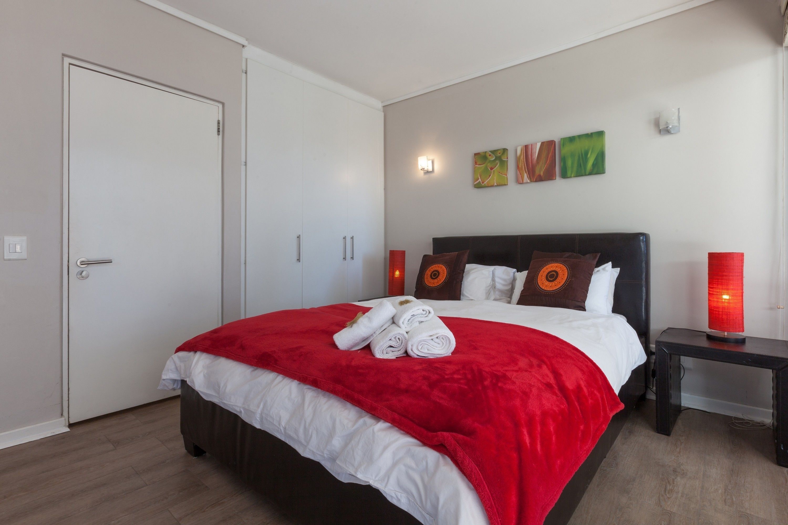 Una cama bien hecha con una manta roja y sábanas blancas, una mesita de noche con una lámpara roja y una puerta a la izquierda.
