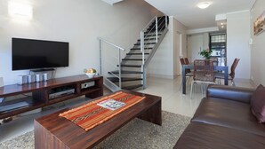 A modern living room with a flat-screen TV, wooden coffee table, and a sofa. A staircase leads to a dining area with a table and chairs. - Adderley Terraces J10 by CTHA (Cape Town)