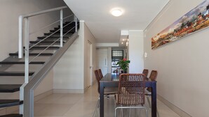 A modern interior with a staircase, a dining area with wicker chairs, and a cityscape painting on the wall. - Adderley Terraces J10 by CTHA (Cape Town)