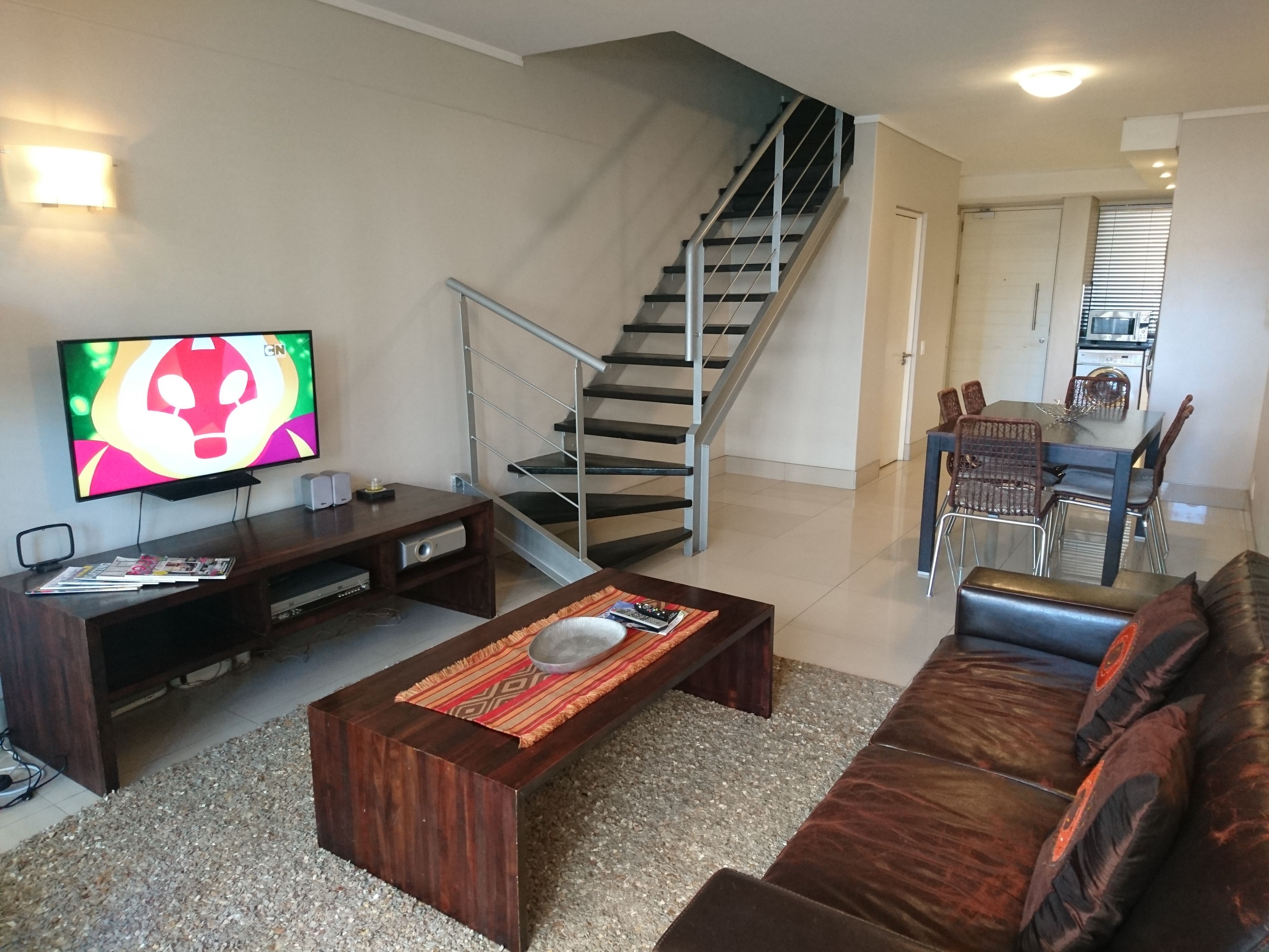 Una sala de estar con sofá, mesa de centro, televisor, mueble de entretenimiento, escalera, comedor y cocina.