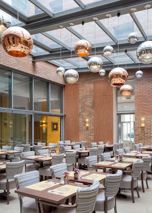 A spacious restaurant with high ceilings, mirrored spherical light fixtures, and a brick wall. Tables are set with striped chairs and centerpieces. - Marmara Hotel Budapest (Budapest)