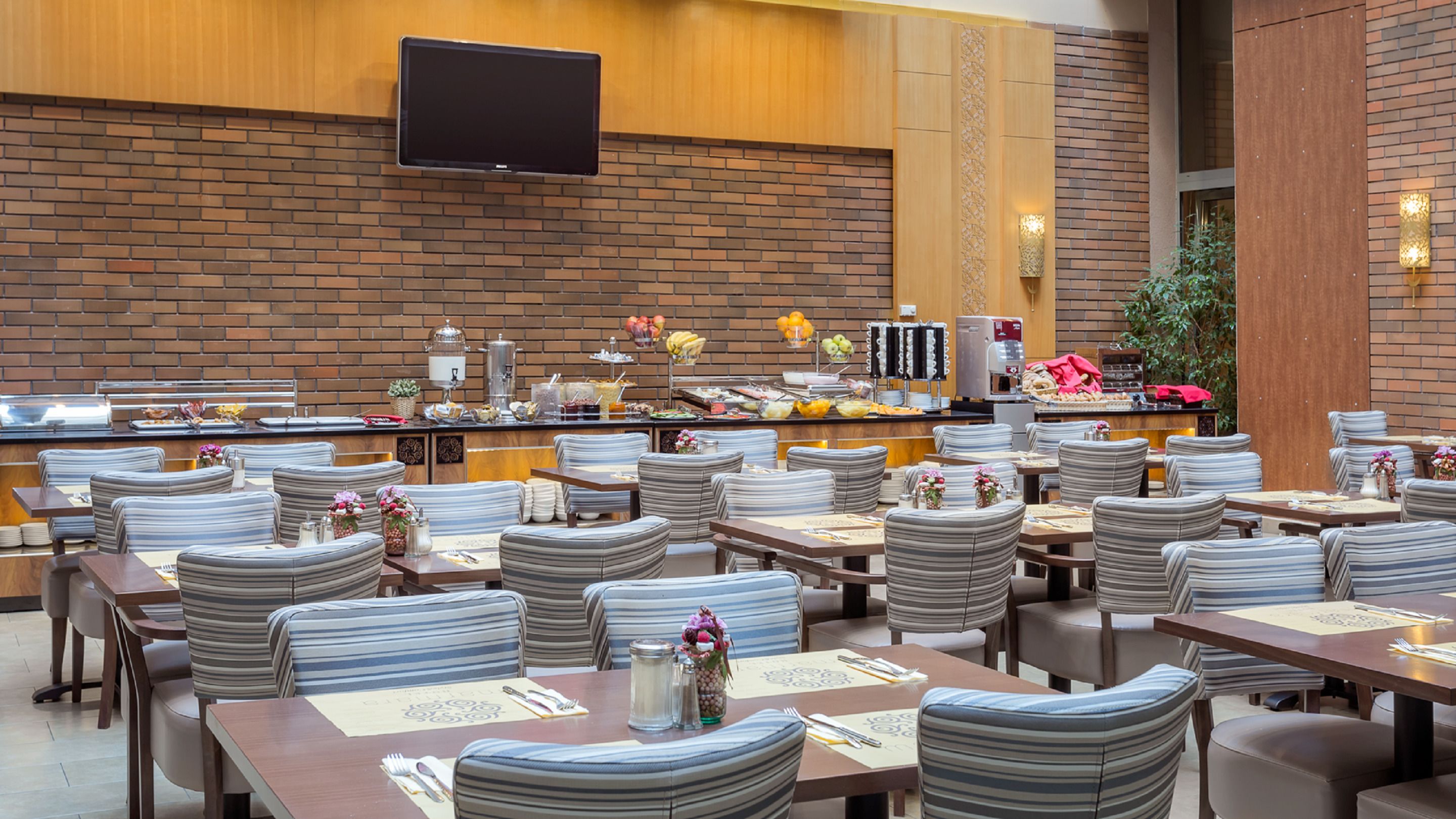 A restaurant with striped chairs and tables, a buffet with various food items, and a flat-screen TV mounted on the wall.
