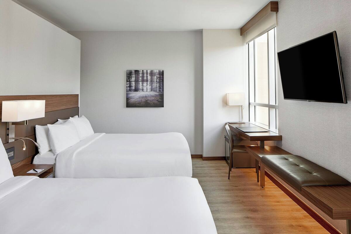 A hotel room with two beds, a desk, a bench, a flat-screen TV, and a painting on the wall.