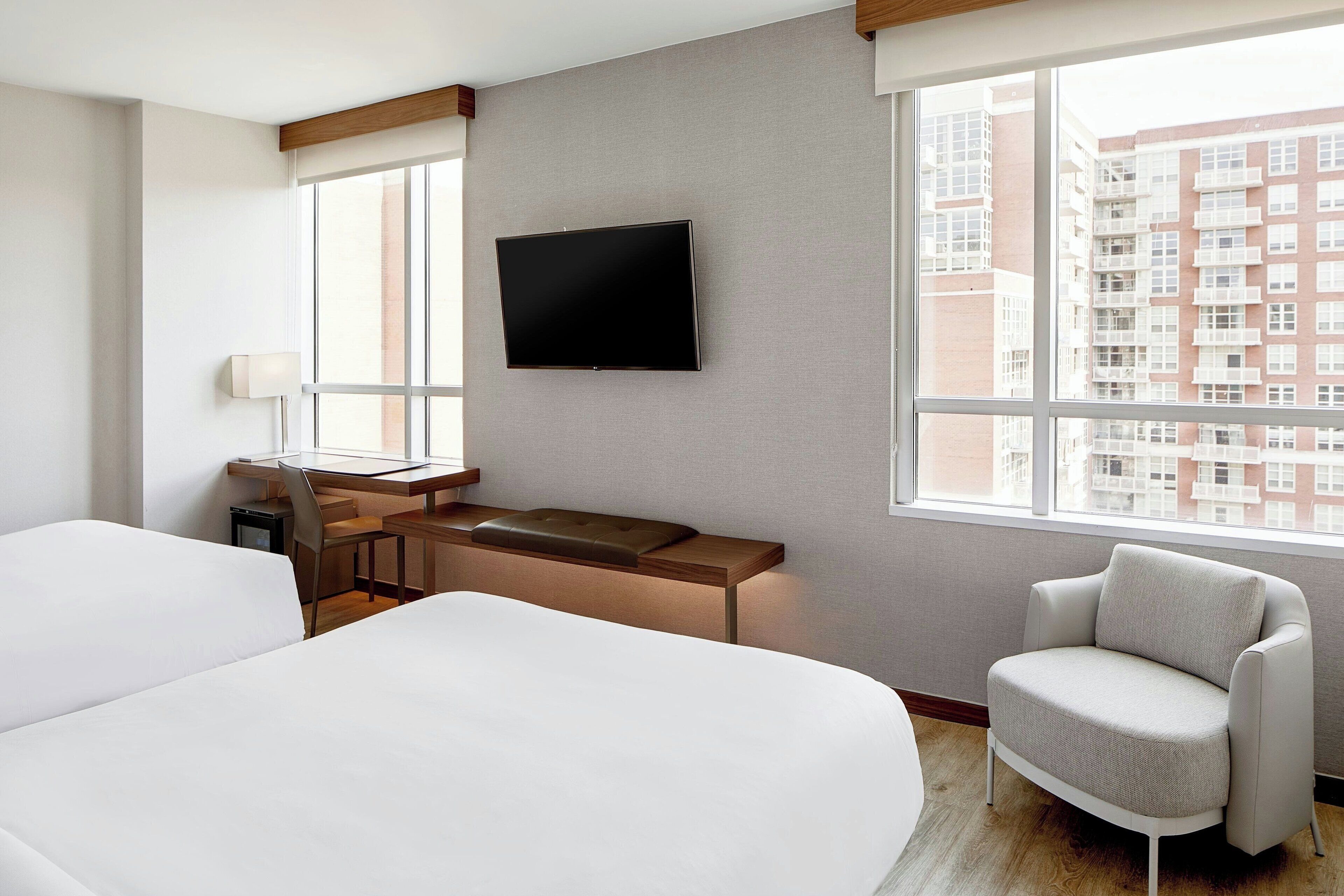 A hotel room with two beds, a desk with a chair, a TV mounted on the wall, and a large window offering a city view.