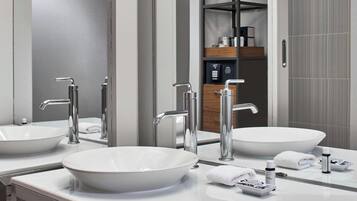 Un baño moderno con dos lavamanos grandes blancos, grifos cromados y un espejo. Hay artículos de aseo en la encimera, y se ve un perchero.