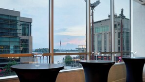 Habitación de hotel moderna con ventanas amplias que ofrecen vistas del horizonte de la ciudad y una masa de agua. Hay tres mesas negras con copas de vino sobre ellas.