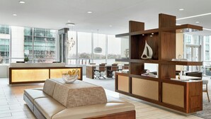 Un moderno vestíbulo de hotel con sofá, mostrador de recepción de madera y grandes ventanales con vistas a la ciudad.