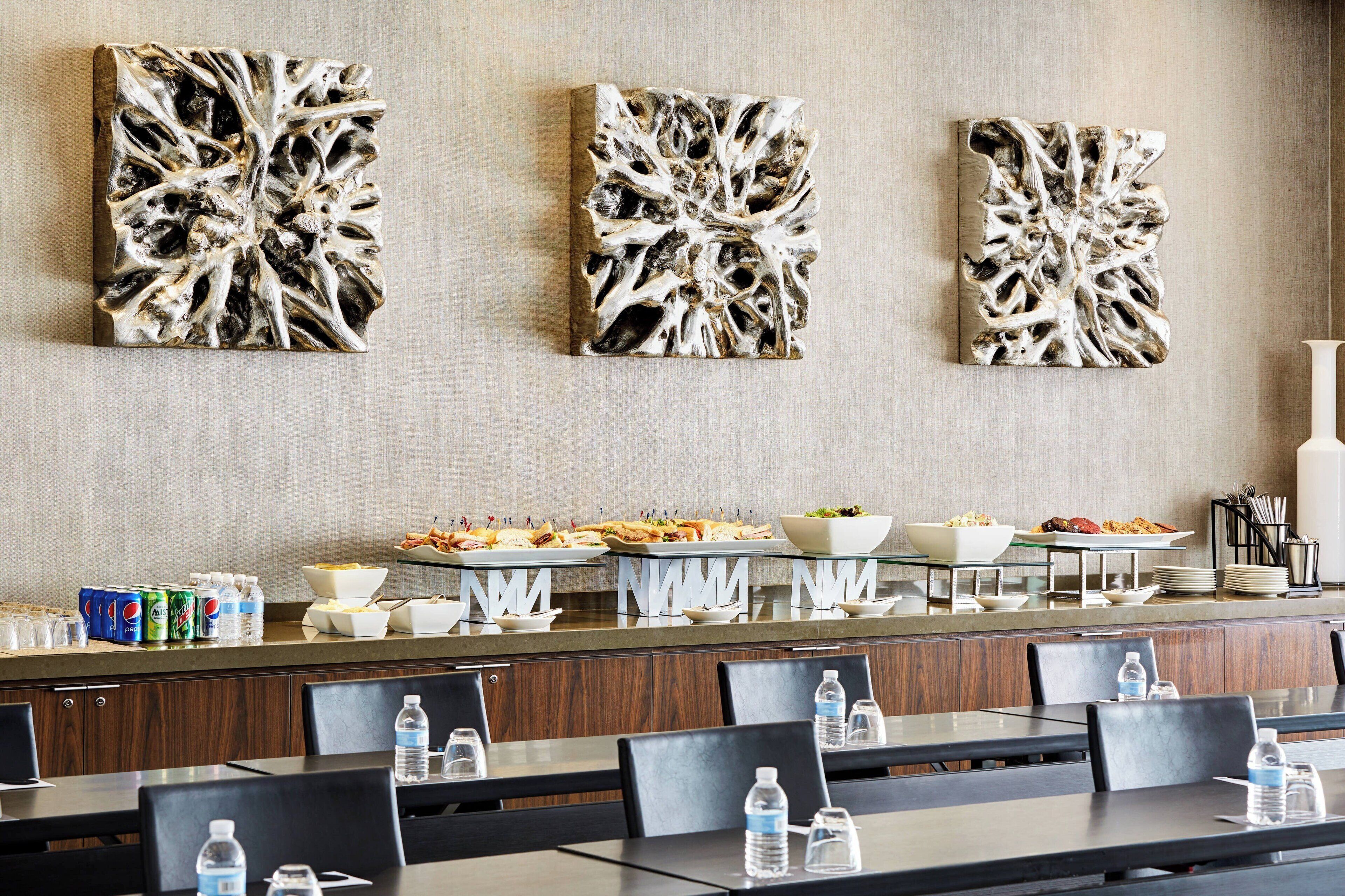 A hotel dining area with three abstract wall art pieces, buffet setup with food and drinks, and neatly arranged tables and chairs.