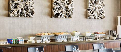 A hotel dining area with three abstract wall art pieces, buffet setup with food and drinks, and neatly arranged tables and chairs.
