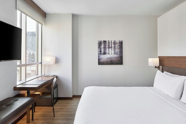 Une chambre d’hôtel équipée d’un lit, d’un bureau, d’une chaise, d’une télévision et d’une fenêtre donnant sur des immeubles.