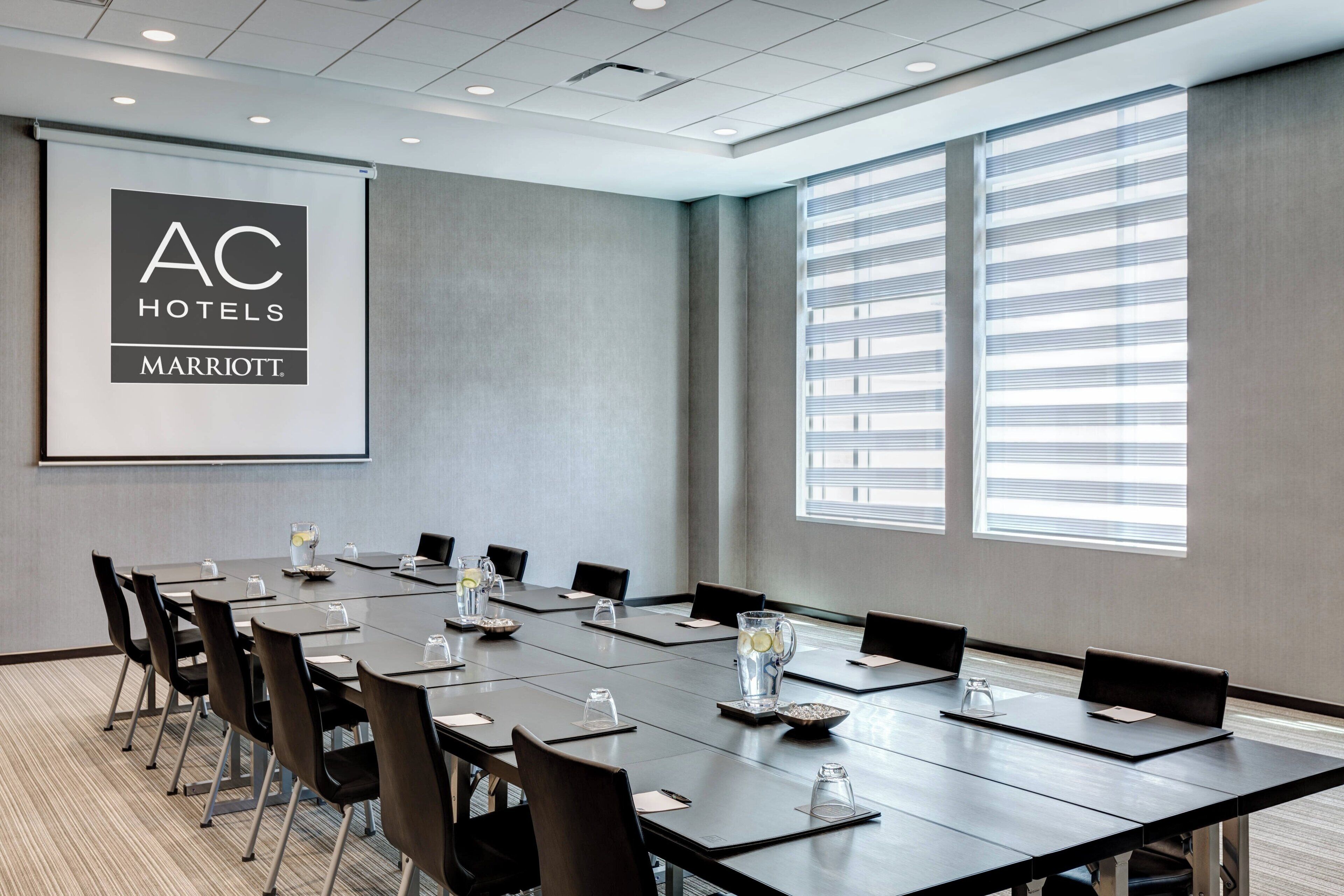 Une salle de conférence dotée d’un grand écran affichant le logo d’AC Hotels Marriott, d’une longue table entourée de chaises et de grandes fenêtres équipées de stores.