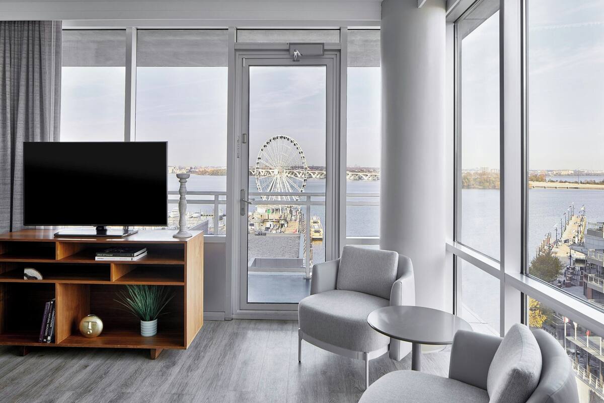 A modern living room with a large TV, a wooden entertainment unit, and a seating area with a view of a Ferris wheel and water.