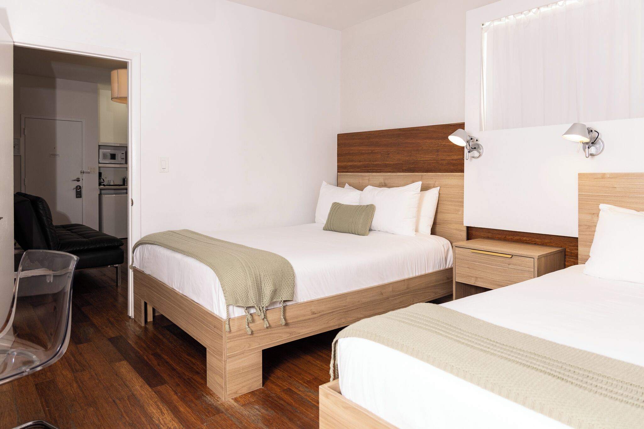 A hotel room with two beds, a wooden headboard, a bedside table, a chair, and a kitchenette in the background.